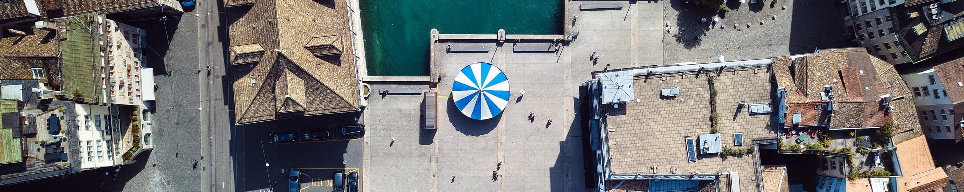 An aerial view of a city square with a blue and white striped canopy at its center, surrounded by historic buildings and a body of water on one side. The area is mostly empty, with a few people visible as small figures.