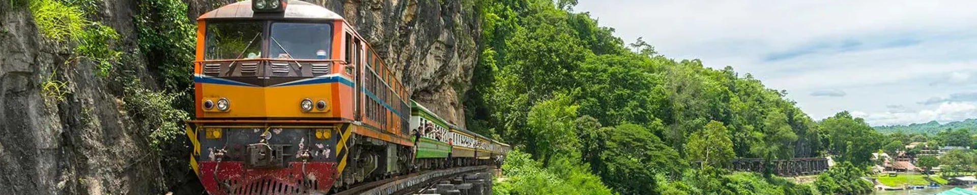 Zug fährt auf einer schmalen Eisenbahnstrecke entlang einer steilen Felswand über einer Flusslandschaft. Der Zug ist orange und bewegt sich auf einer Holzbrücke, während sich rechts ein grünes Tal mit Bäumen und einem Fluss unter blauem Himmel erstreckt.