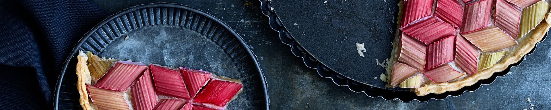 Rhubarb tart decorated with pink and green rhubarb pieces arranged in a geometric pattern, partially cut. One slice sits on a plate beside the tart, with milk and sugar on a dark surface.