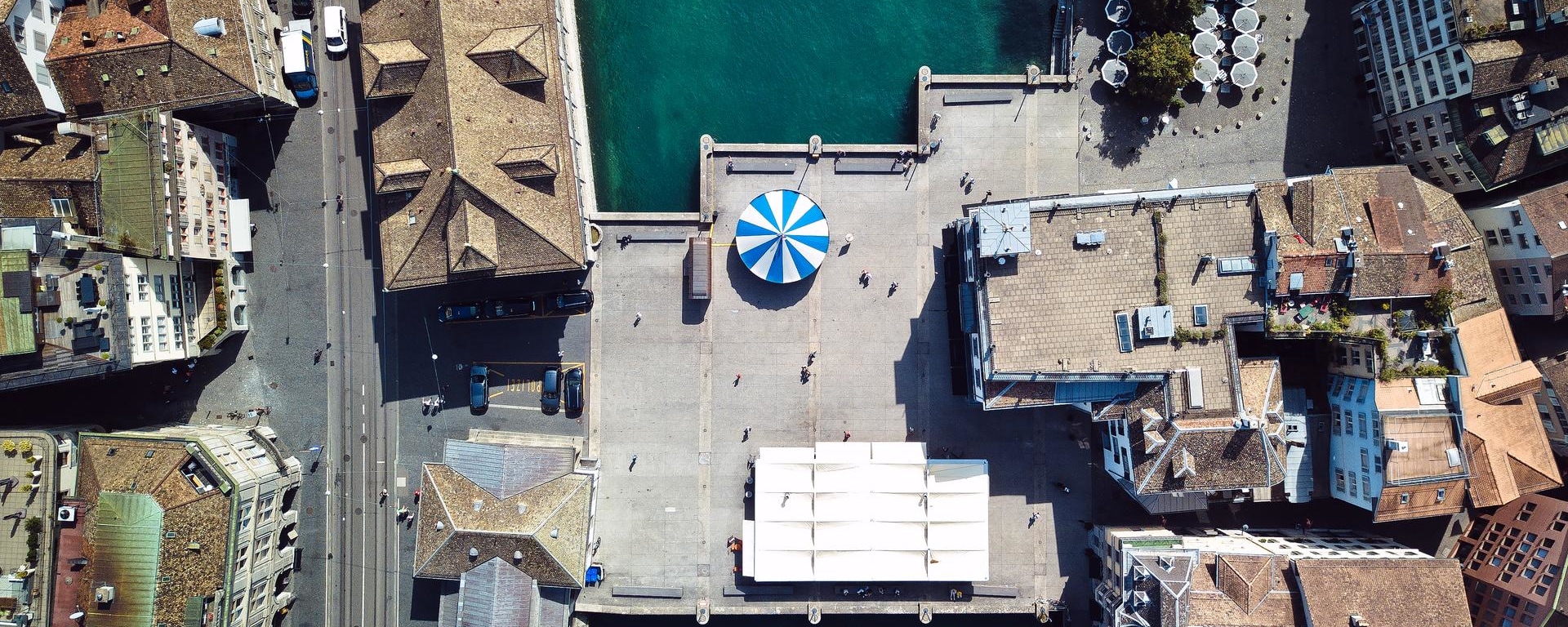 An aerial view of a city square with a blue and white striped canopy at its center, surrounded by historic buildings and a body of water on one side. The area is mostly empty, with a few people visible as small figures.