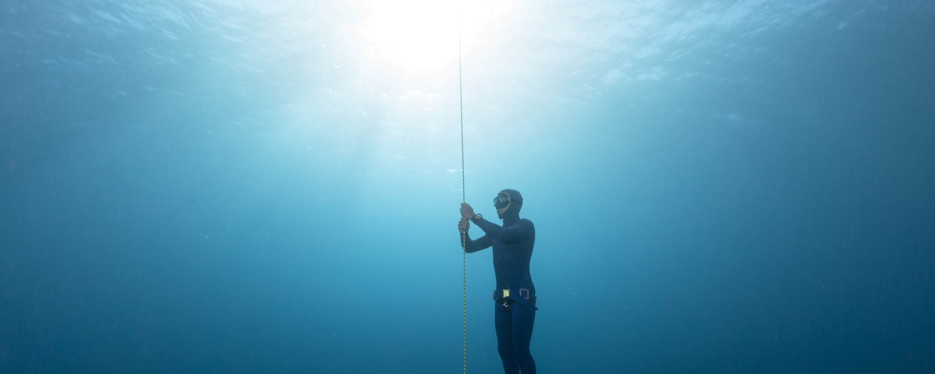 Eine Person im Neoprenanzug und mit Tauchmaske steigt unter Wasser an einem vertikal verlaufenden Seil in klarem, blauem Wasser auf.