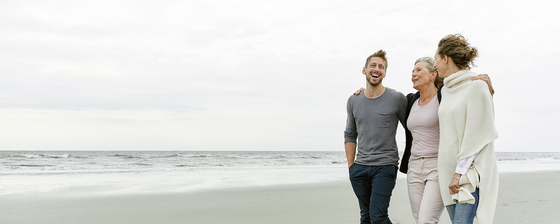 An older women has her arms around a young man and a young woman while walking along a beach