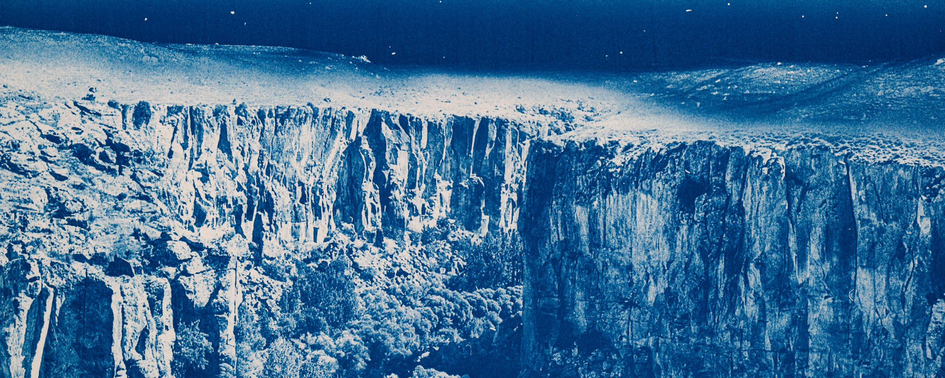 Blue-toned image of steep rocky cliffs and rugged landscape under a dark sky, with textured terrain extending across the horizon.