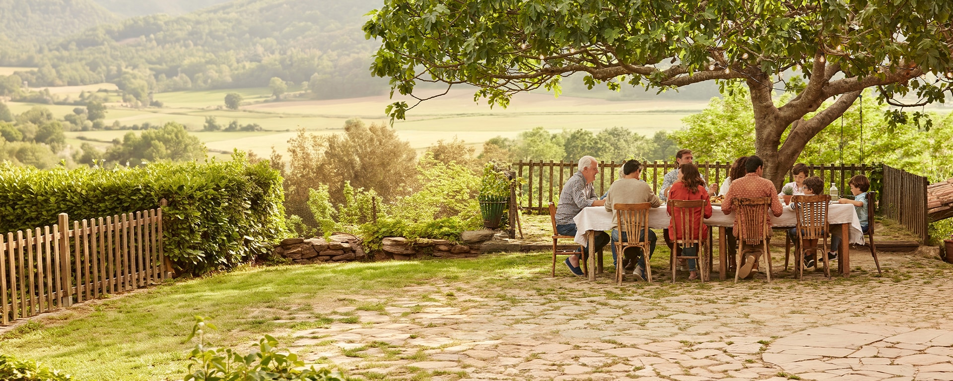 People seated around a table beneath a large tree in a garden overlooking a rural countryside landscape.