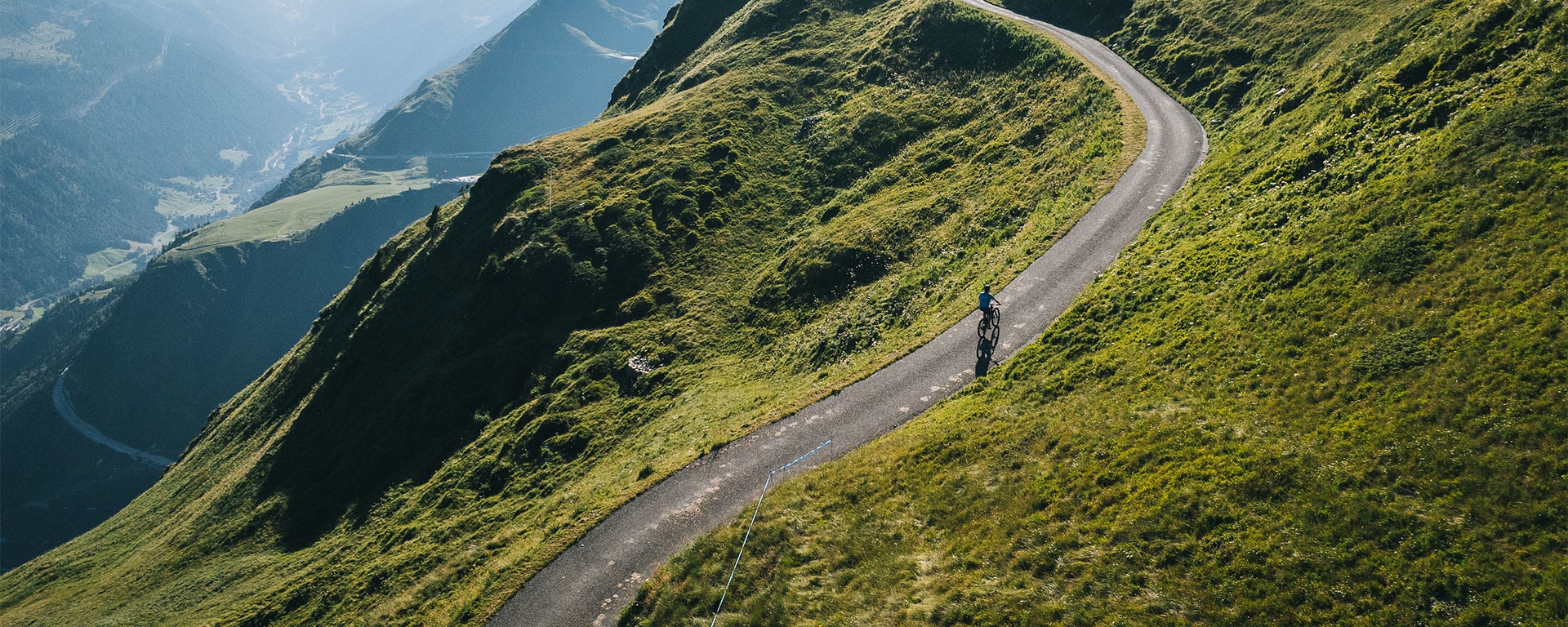 Radfahrer auf einer kurvenreichen Bergstrasse in den Schweizer Alpen unter sonnigem Himmel - atemberaubende Aussicht auf grüne Hänge und ferne Alpengipfel.