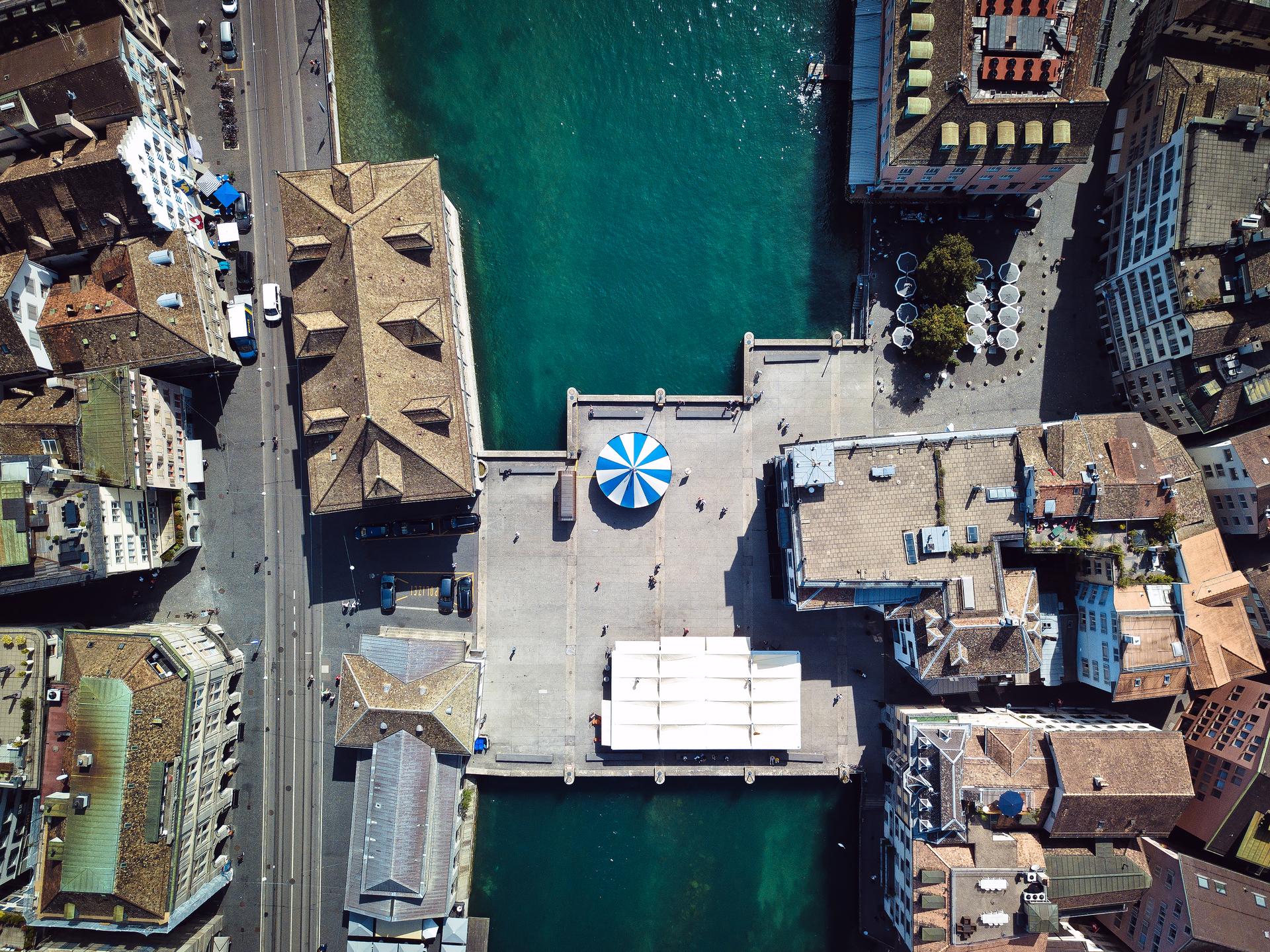 Eine Luftaufnahme eines Stadtplatzes mit einem blau-weiss gestreiften Baldachin in der Mitte, umgeben von historischen Gebäuden und einem Gewässer auf einer Seite. Der Platz ist grösstenteils leer, mit einigen wenigen Personen, die als kleine Figuren zu sehen sind.
