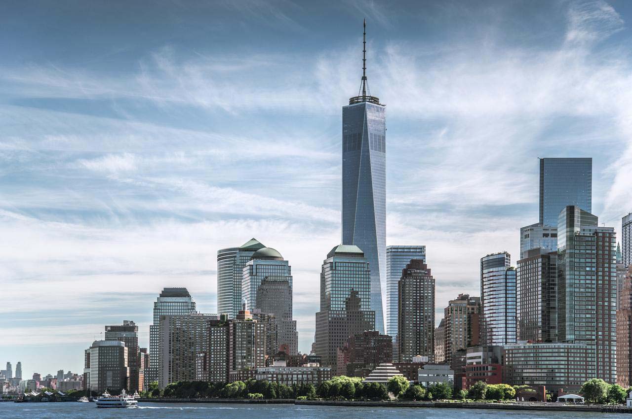 Photo of the New York skyline. Several tall commercial buildings stand near to the shore, which is lined with trees.