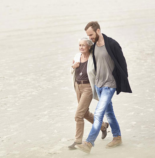 Man with one arm over his mother's shoulder and walking with her at the beach.