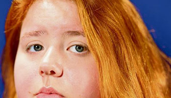 A young woman with red hair and blue eyes, looking directly at the camera with a confident expression.
