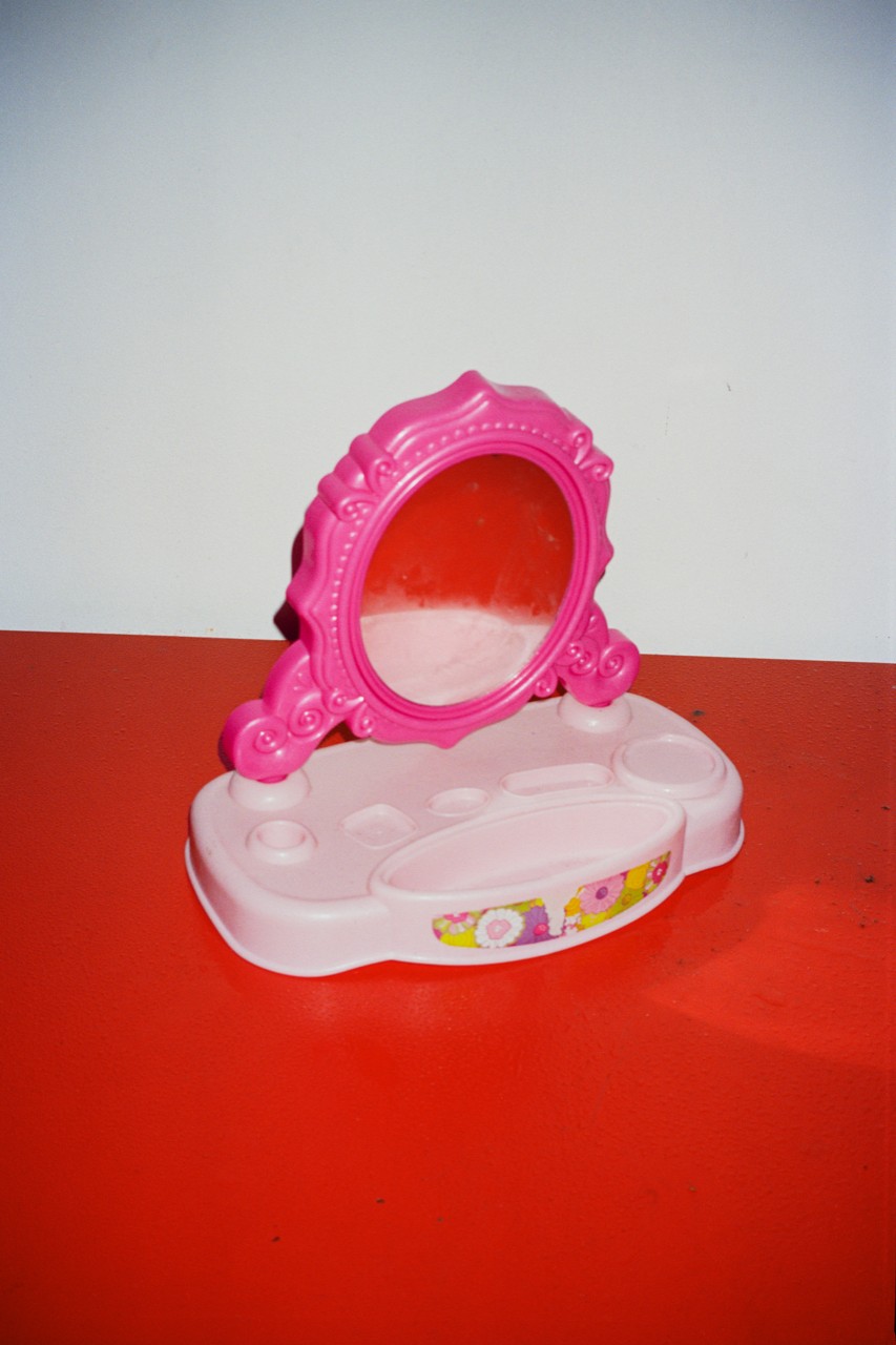 Small plastic toy vanity with a pink oval mirror and a white base placed on a red surface. The background is plain and light, with the object centered in the image.