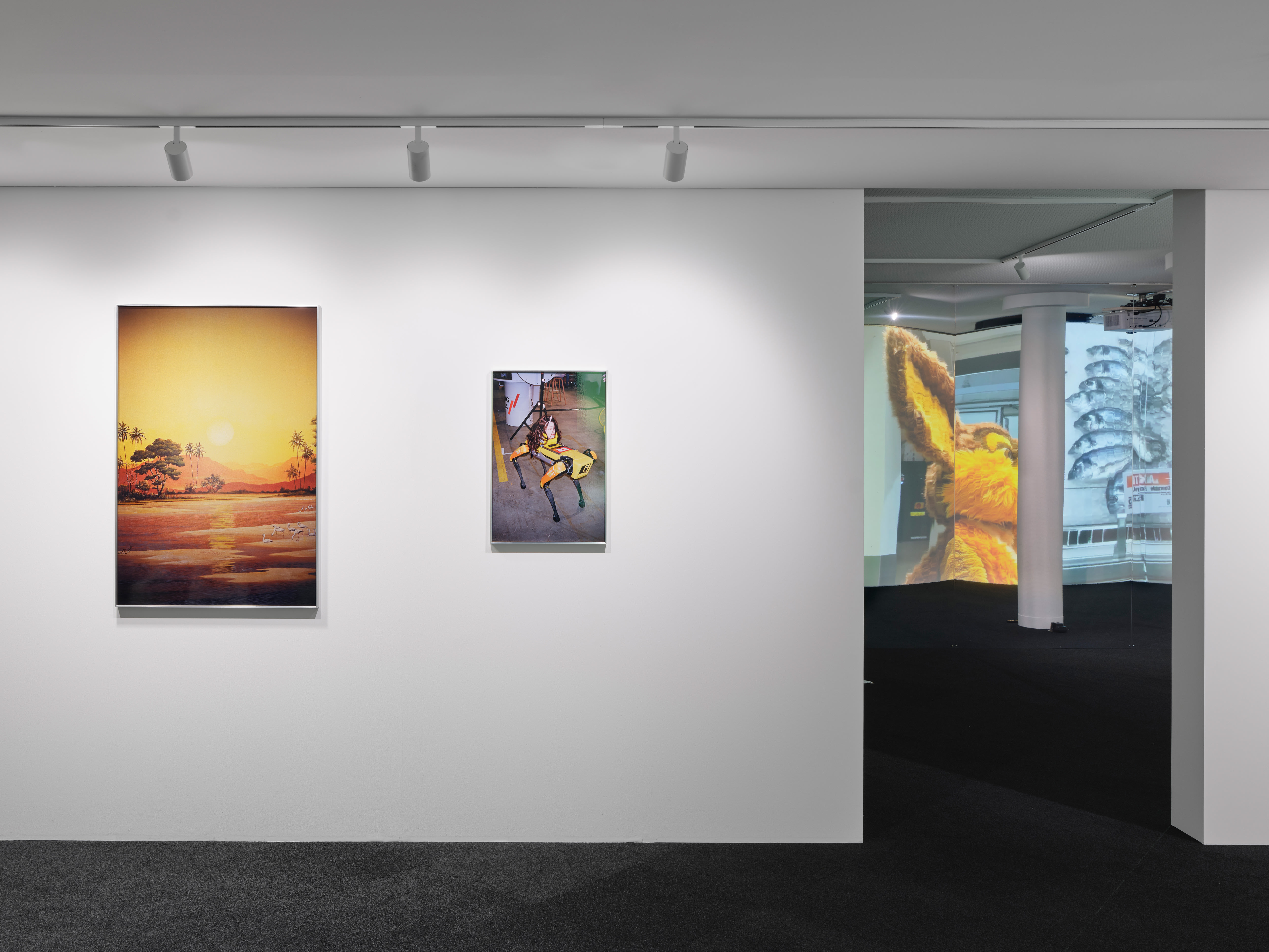 Weisser Ausstellungsraum mit zwei gerahmten Bildern – eine Landschaft bei Sonnenuntergang und eine Szene mit einem Roboterhund – sowie einer Videoprojektion in einem angrenzenden Raum