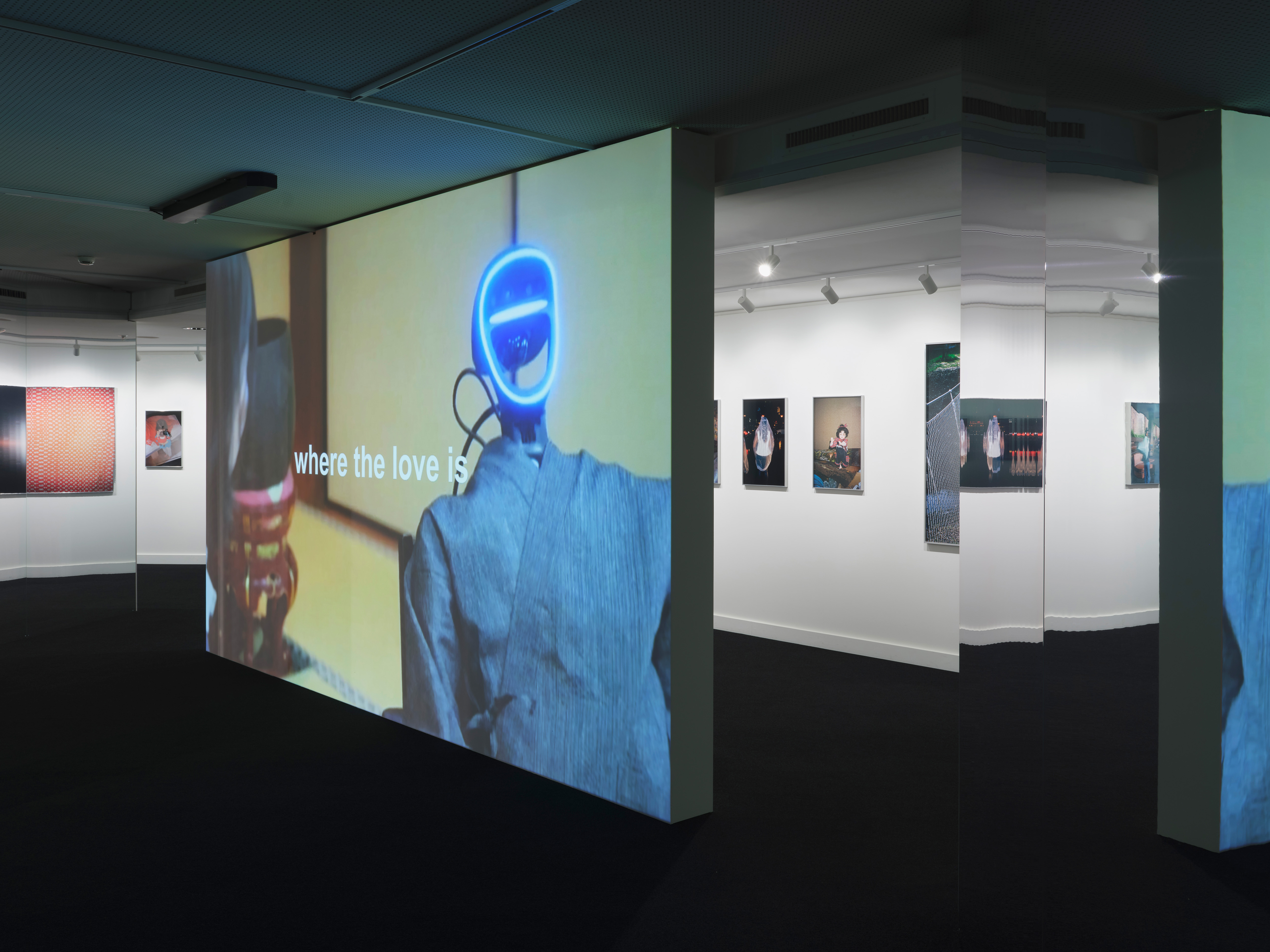 Immersive Videoinstallation in einem Ausstellungsraum mit Projektion einer Figur mit leuchtendem Gesicht und dem Text „where the love is“ auf einer wandfüllenden Fläche