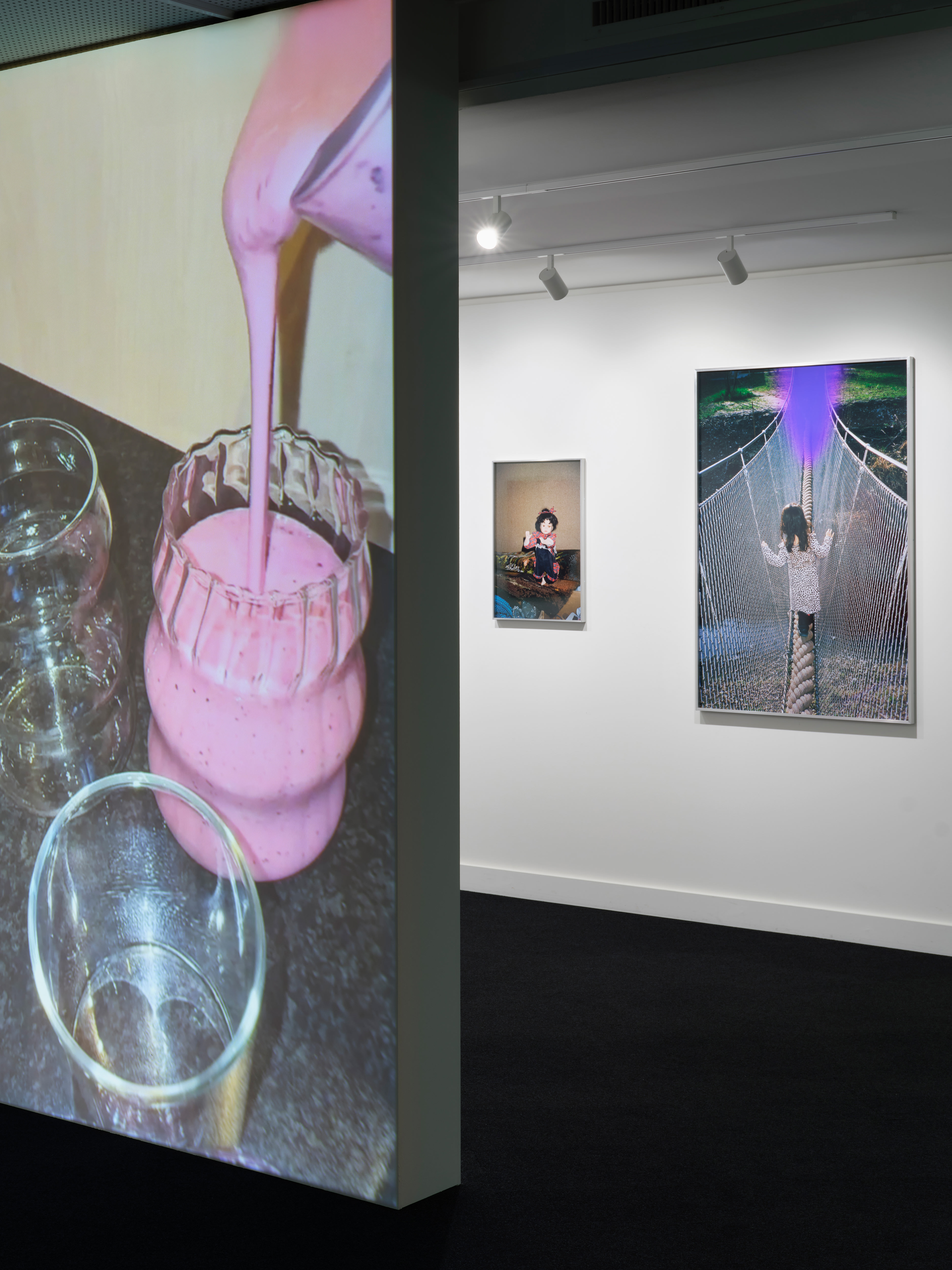 Detail einer Projektion mit rosa Flüssigkeit, die in ein Gefäß gegossen wird, neben Fotografien an einer weißen Ausstellungswand, darunter ein Bild einer Person auf einer Seilbrücke