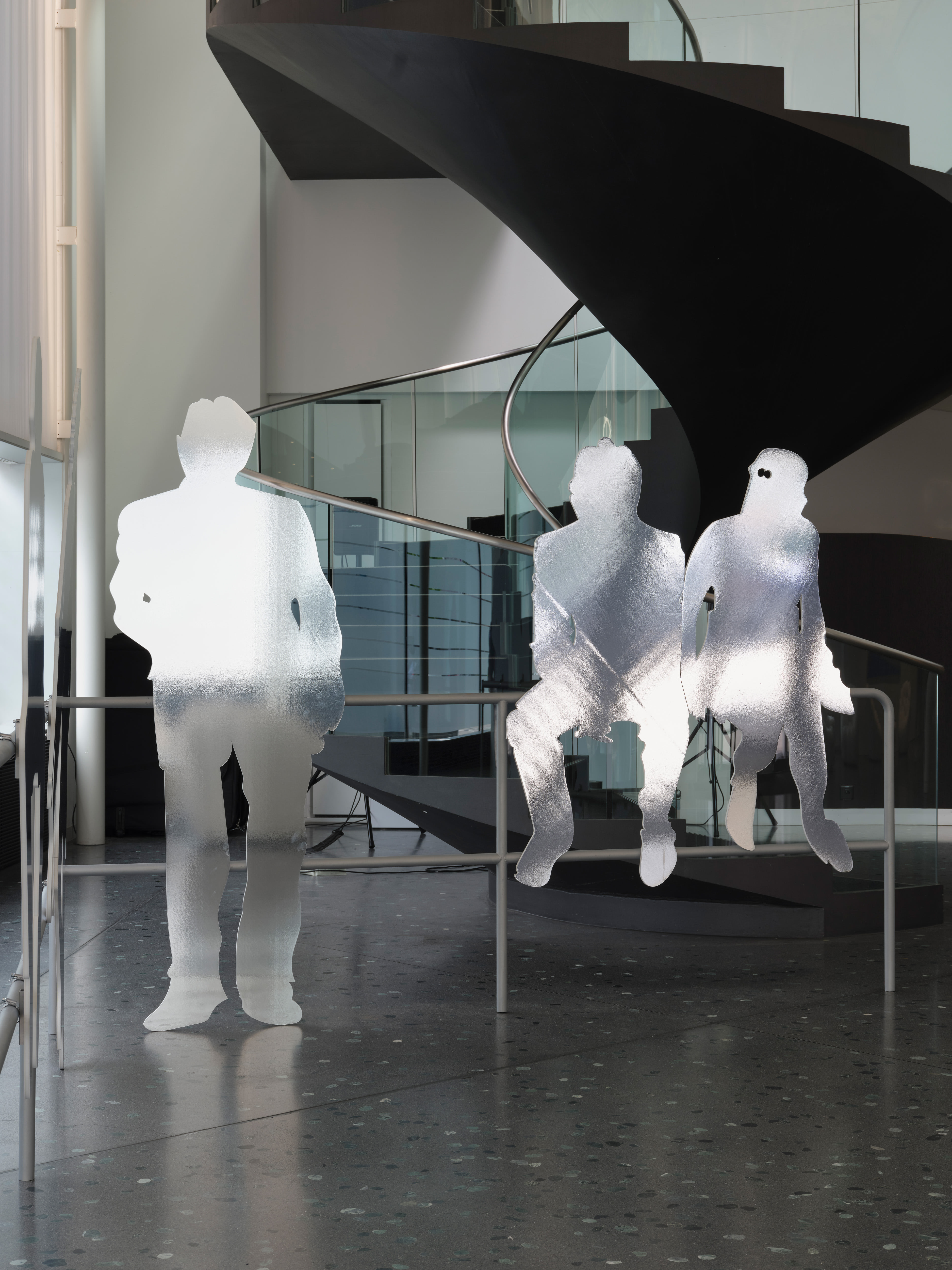 Installation mit transparenten Silhouetten dreier menschlicher Figuren vor einer geschwungenen Treppe in einem modernen Innenraum