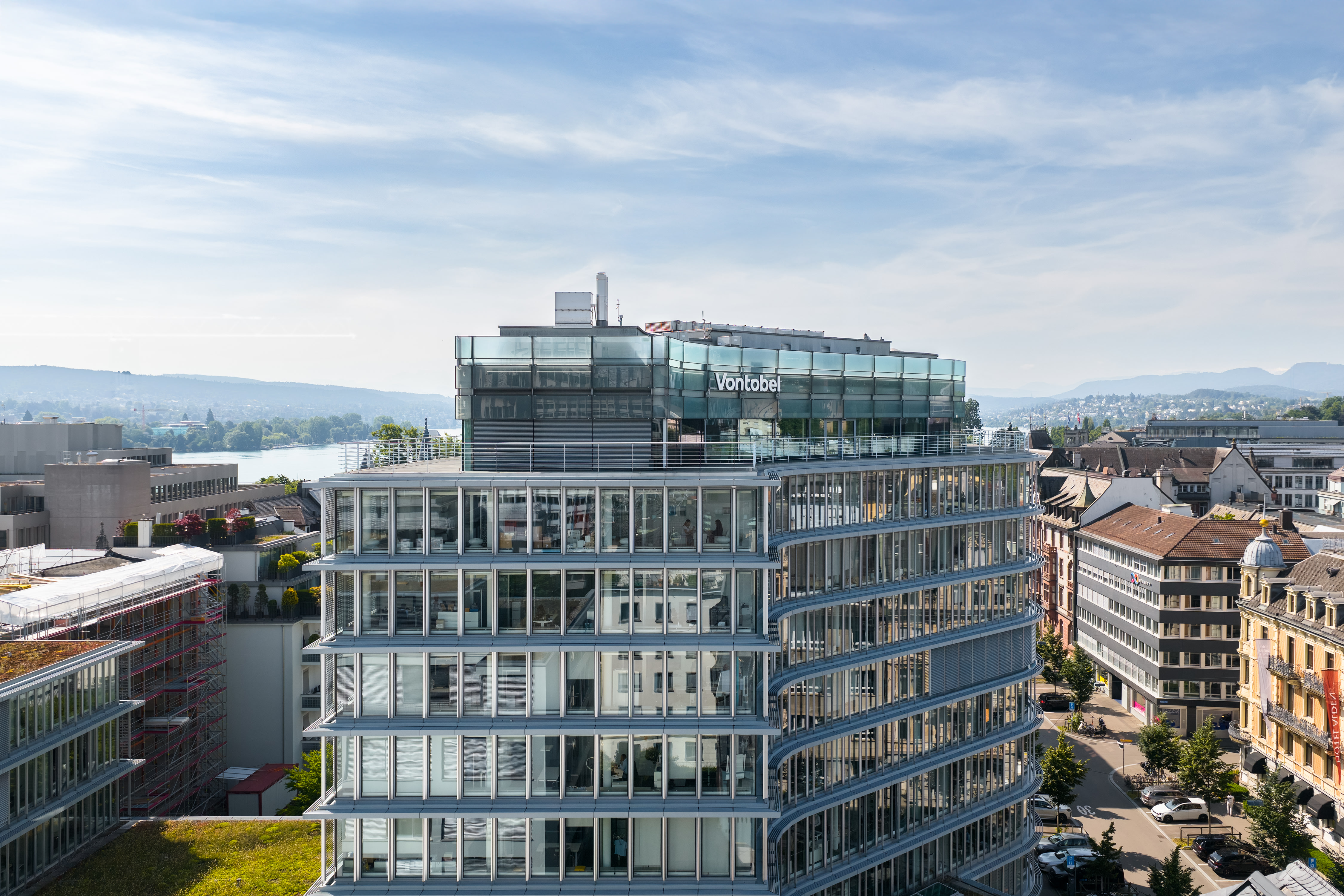 Modern glass office building with “Vontobel” signage in Zurich, overlooking the city and a river under a blue sky.
