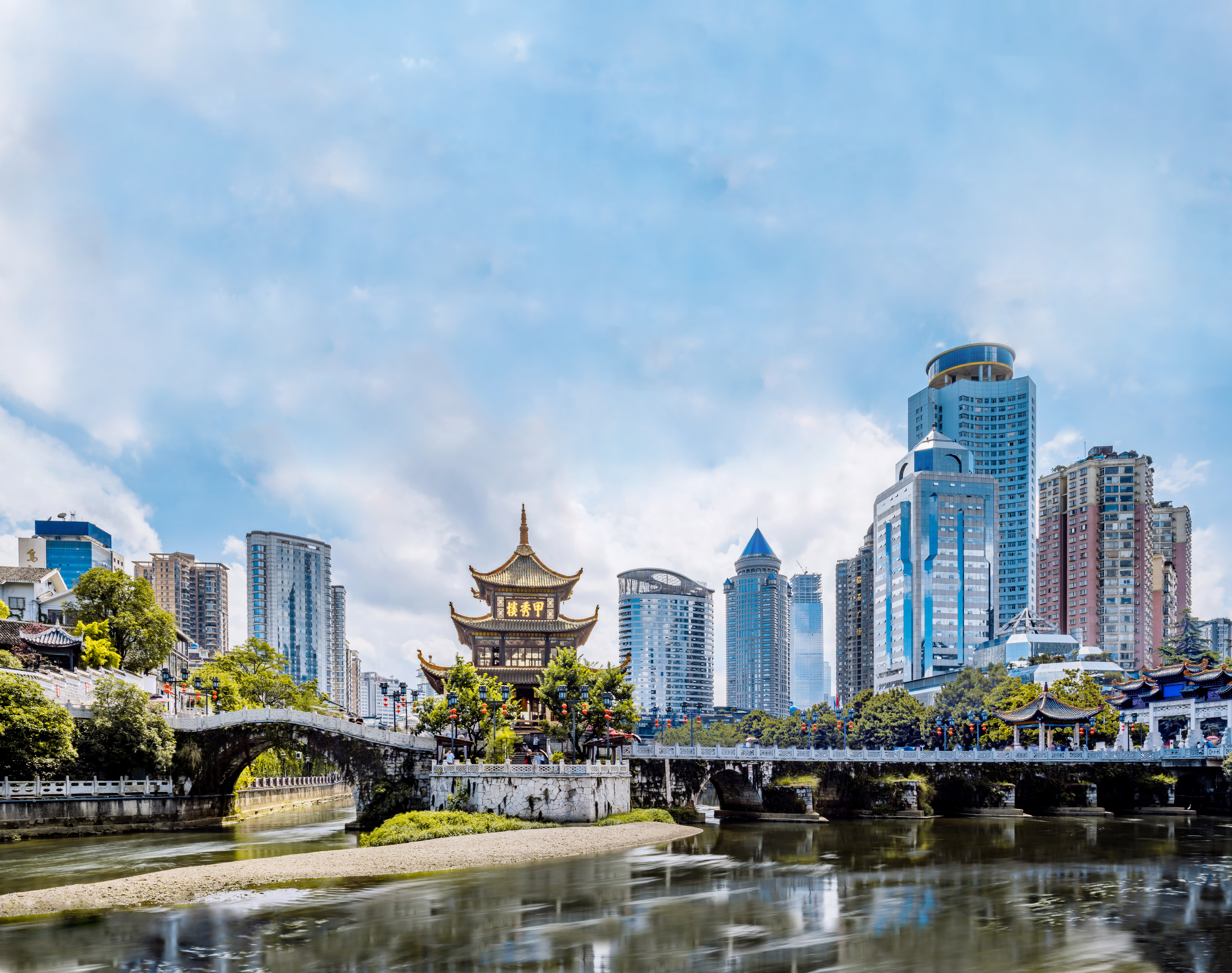Stone bridge with ornate details and flowers over a calm river, traditional pavilion and modern skyscrapers behind
