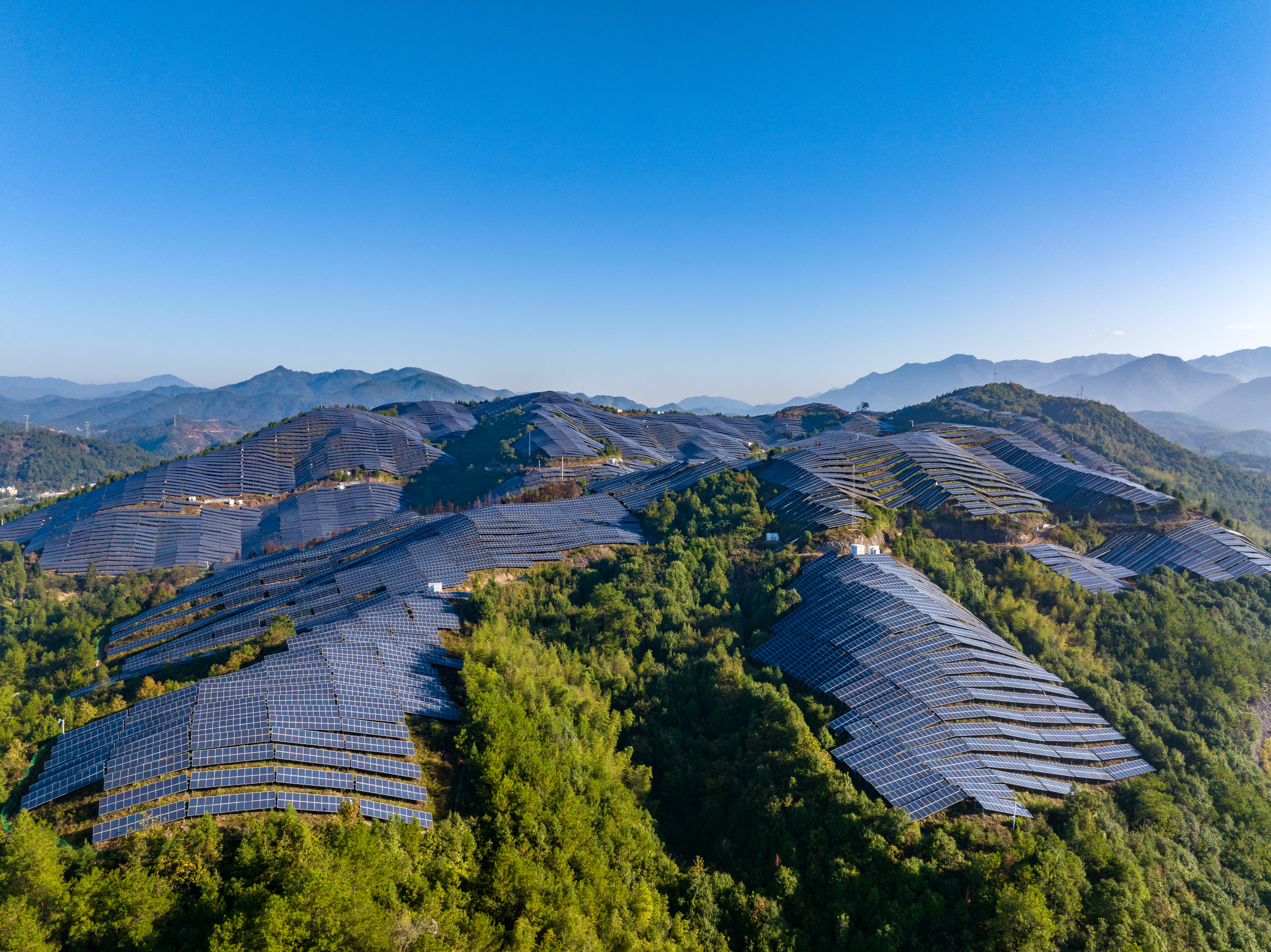 Rows of solar panels form wave-like arrays across green rolling hills, with distant mountains under a clear blue sky.