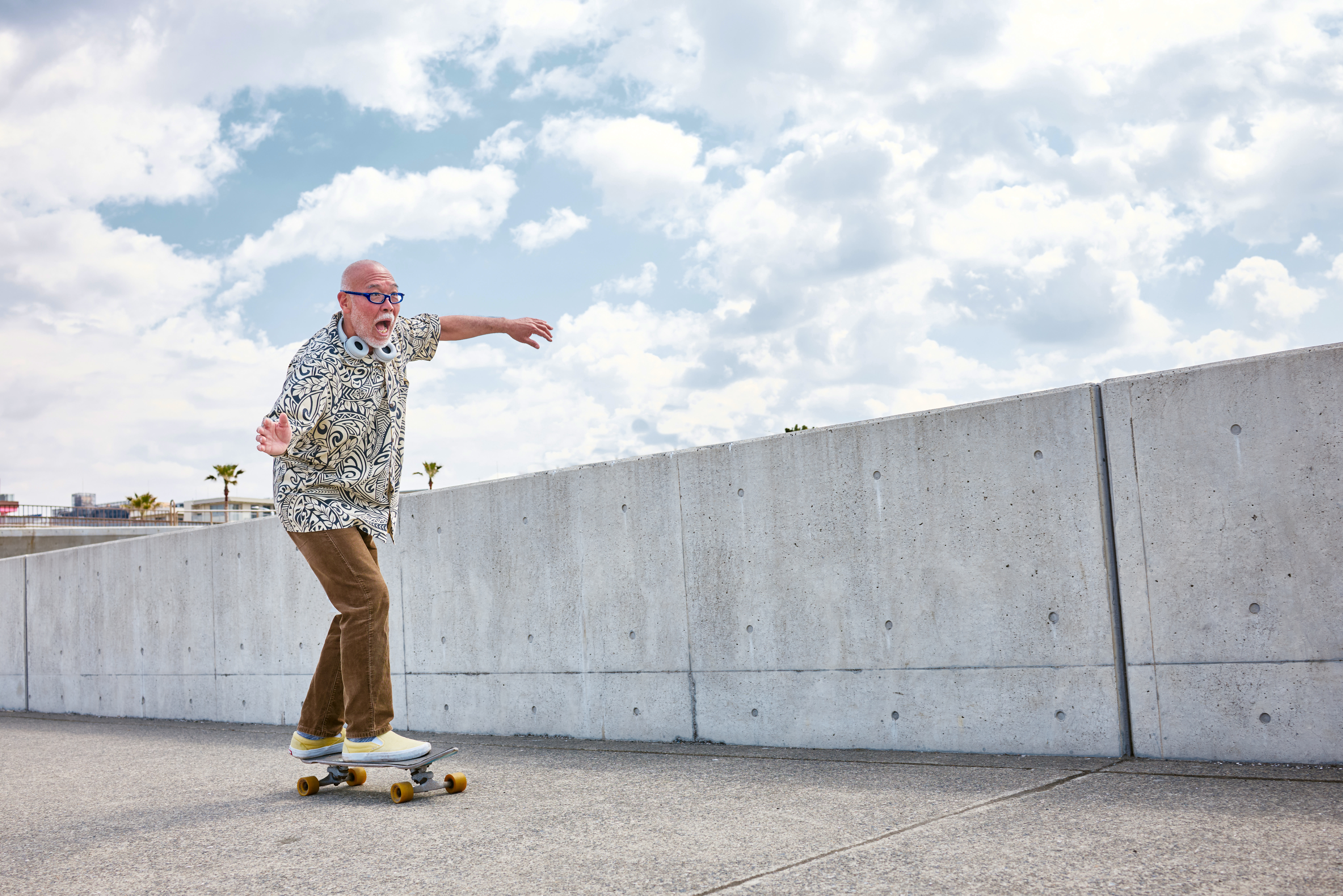 A person skateboarding on a sunny palm-lined boardwalk wearing a patterned shirt, brown pants, skateboard with yellow wheels