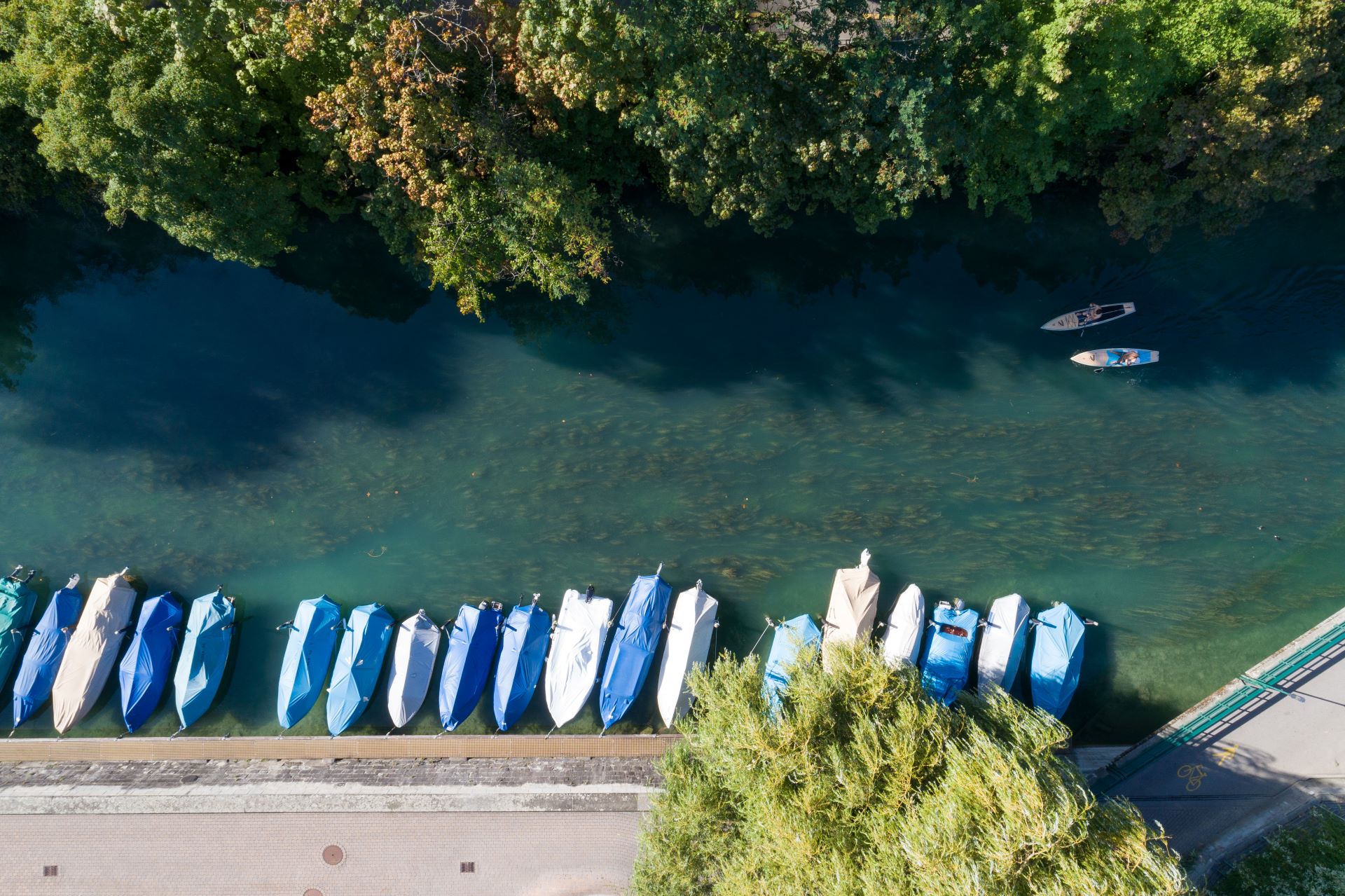 Aerial view of a calm riverbank with moored boats covered in blue and beige tarpaulin, alongside a paved walkway. Two individuals are seen stand-up paddling on the water, with lush trees lining the river's edge.