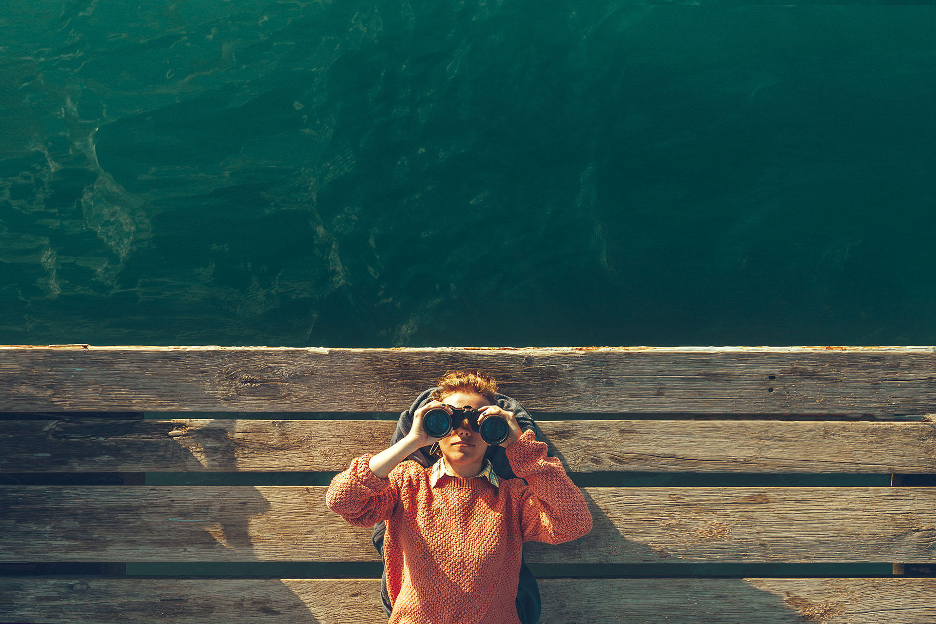 Top view of a person on a wooden pier, looking through binoculars, with clear green water below.