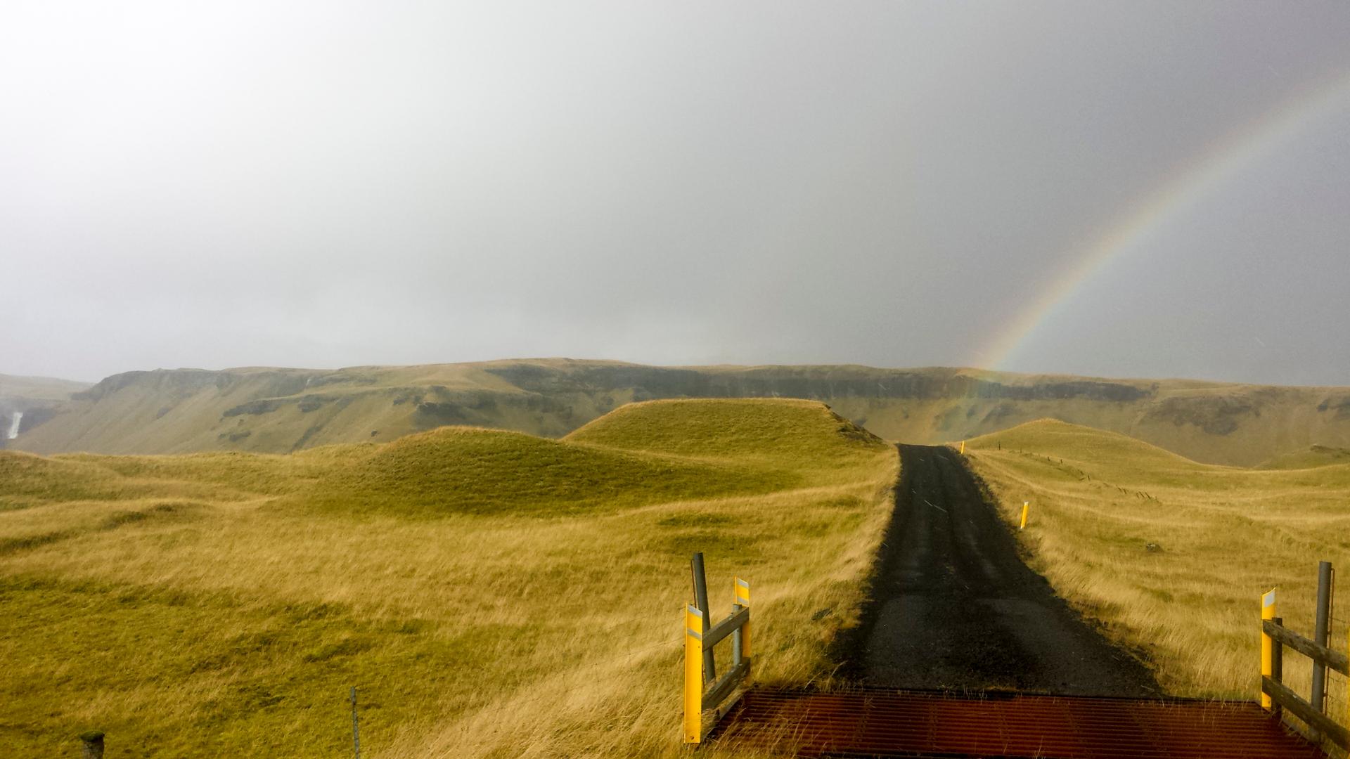 A path and small bridge through a moss and meadow field with a rainbow in the sky
