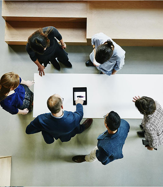 Bird's eye view of a table on which a tablet is lying. Six people are standing around the table.