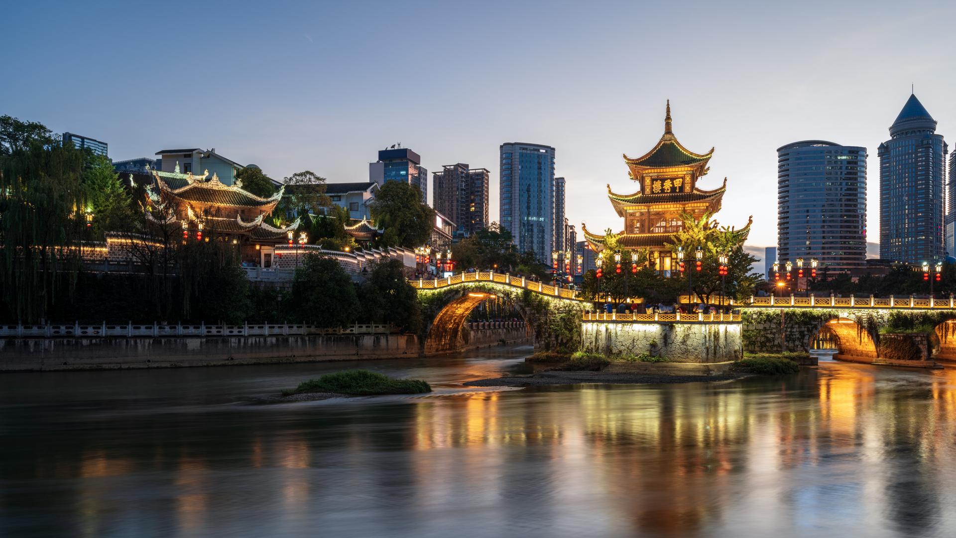 A pagoda and modern buildings line a riverside at dusk with illuminated reflections in the water.