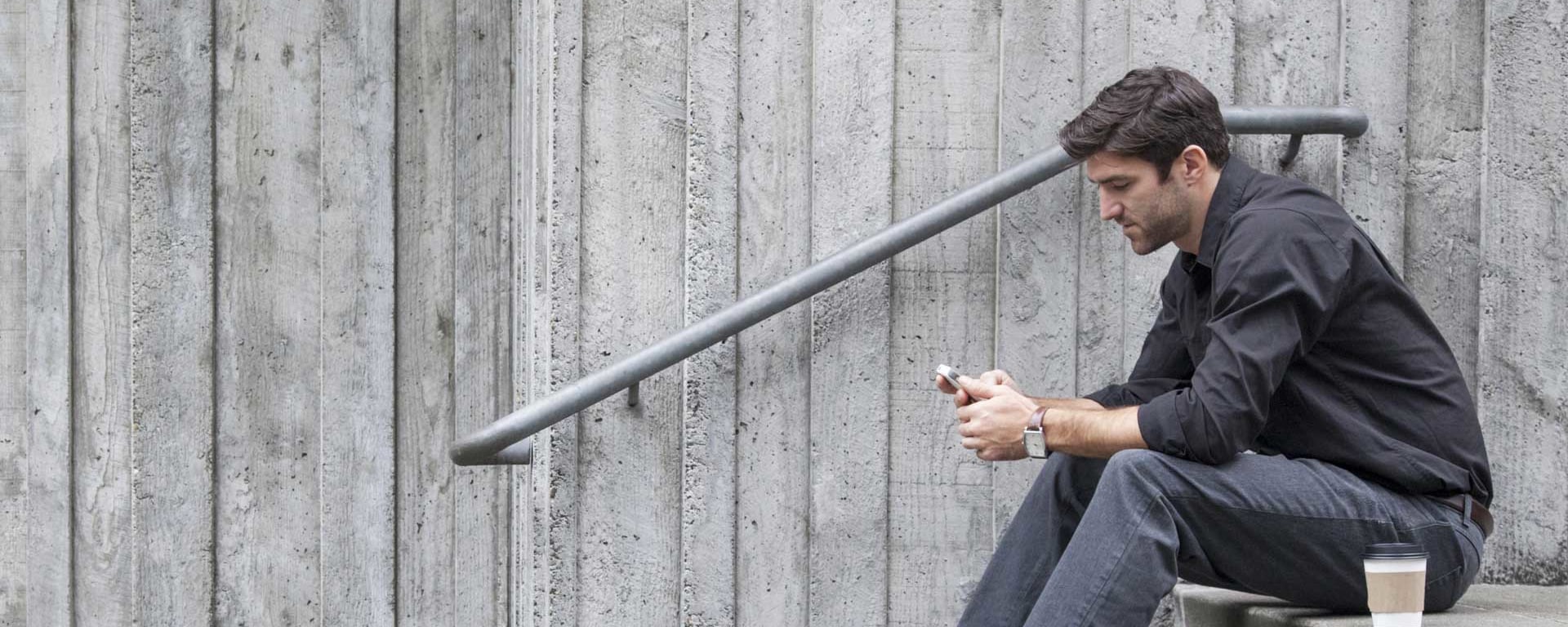 Ein junger Mann mit schwarzer Jacke sitzt auf einer Treppe und schaut in sein Smartphone.