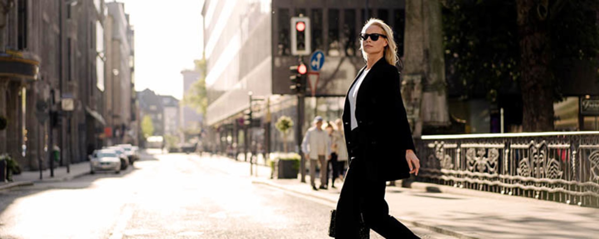 Une femme en tailleur noir et lunettes de soleil traverse une rue avec assurance.