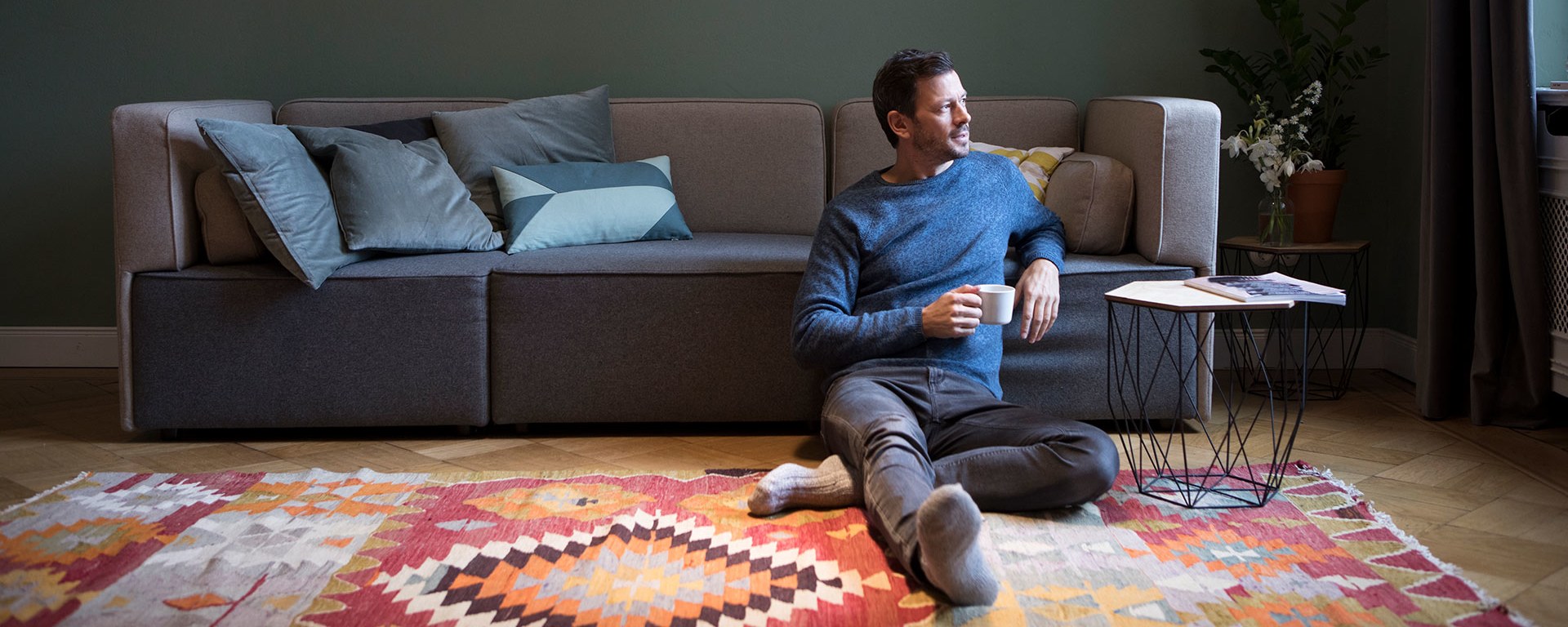 A relaxed man sits on the floor next to a couch, leaning back with a cup in hand, looking to the side. A cozy living room setting with cushions on the sofa.