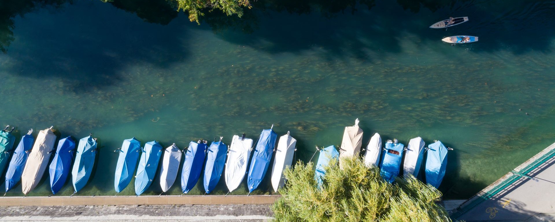 Luftansicht eines ruhigen Flussufers mit angelegten Booten, die mit blauem und beigem Planen bedeckt sind, neben einem gepflasterten Gehweg. Zwei Personen sind auf dem Wasser beim Stand-Up Paddeln zu sehen, mit üppigen Bäumen am Ufer.