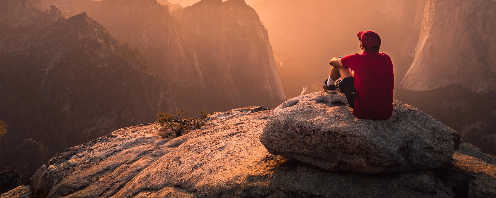 A man with a red cap sits on a rocky ledge and watches the sunset.