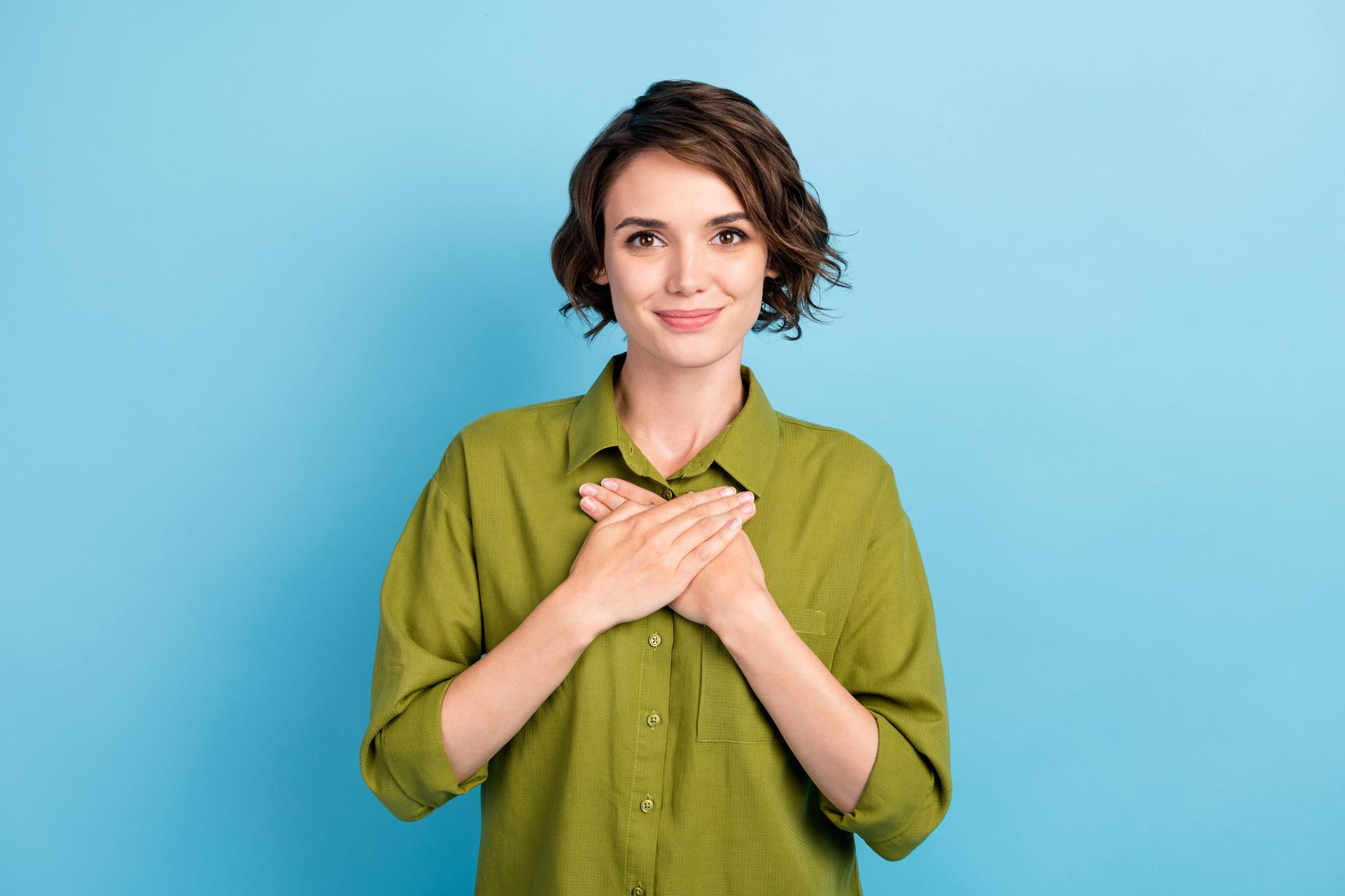A woman in a green shirt with her hands on her chest and a blue background.