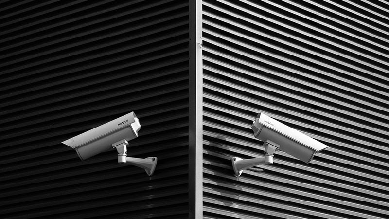 Two white surveillance cameras are mounted on a dark wall and monitor what is happening.