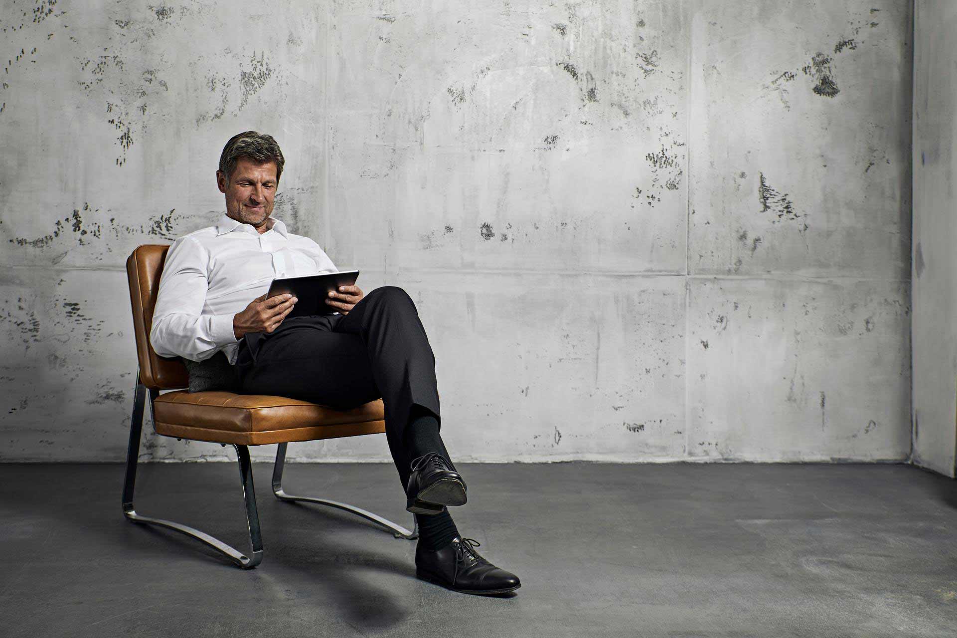 A businessman sits in a leather chair and looks into a tablet.