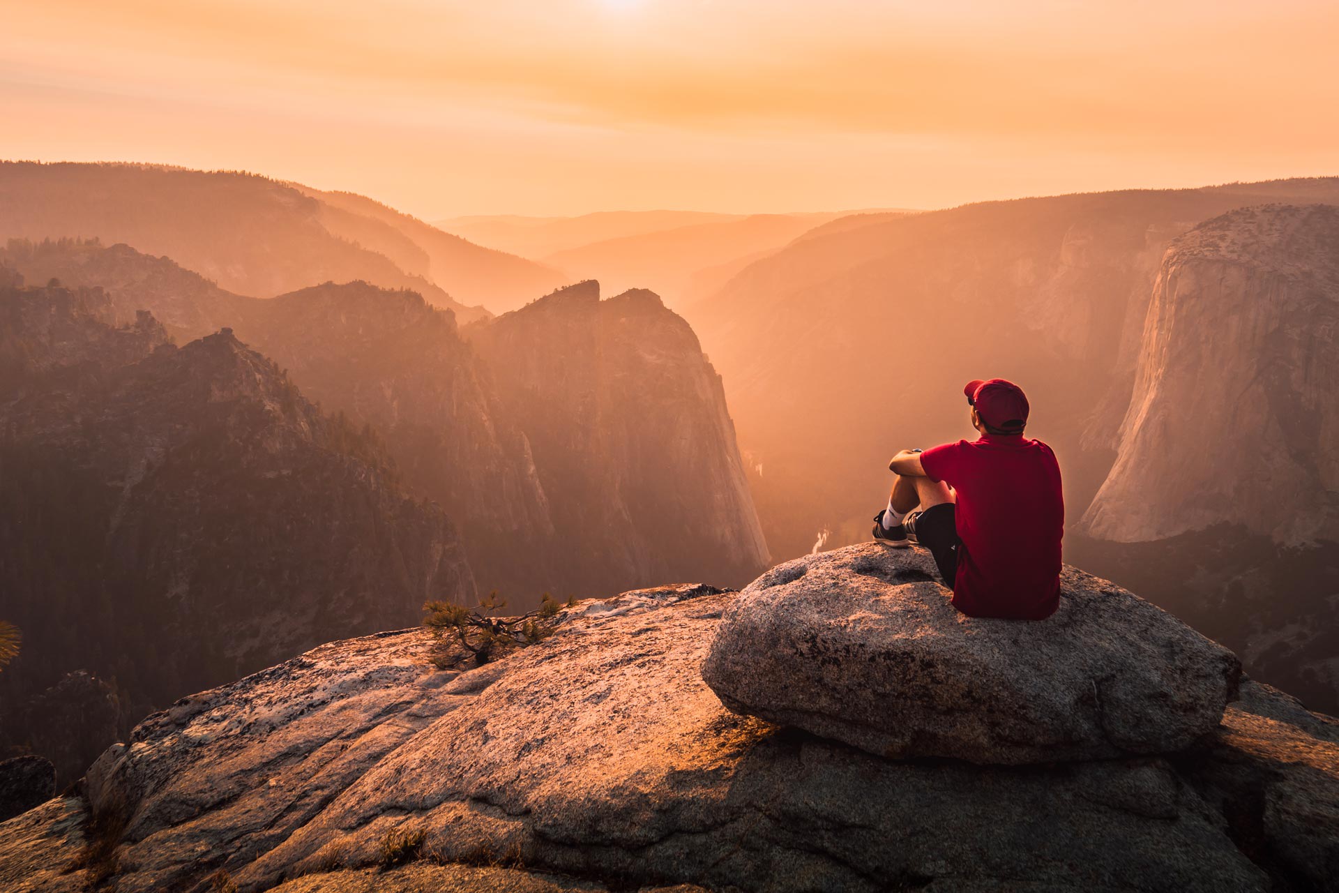Ein Mann mit roter Kappe sitzt auf einem Felsvorsprung und schaut sich den Sonnenuntergang an.