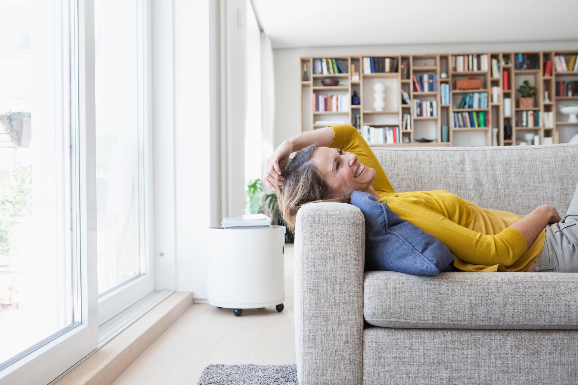 A woman in a yellow sweater is lying on a sofa laughing and enjoying her life insurance.